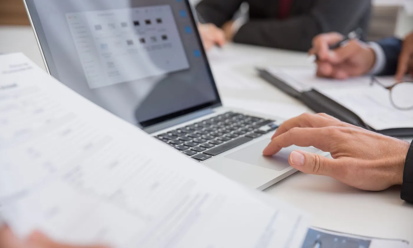 A buyer reviewing printed specifications beside a laptop showing a page of thumbnails