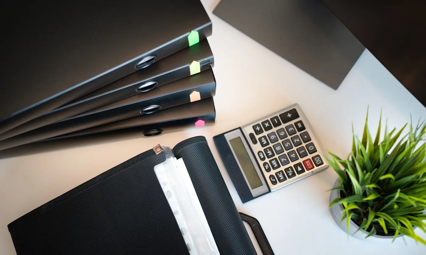 A stack of black office binders with coloured tabs beside a project file, a calculator and a desk plant